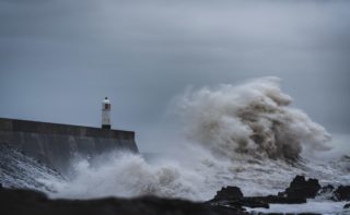 Research: Eine große Welle trifft mit voller Wucht auf eine Kaimauer mit Leuchtturm bei Sturm.